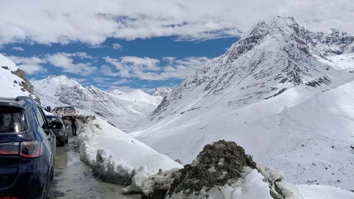 a snowy mountain range with some cars on road in Rohtang Pass road